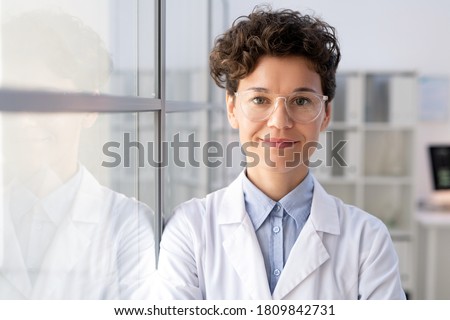 Young successful female worker of scientific laboratory in whitecoat and eyeglasses standing by glass wall inside office in front of camera Royalty-Free Stock Photo #1809842731