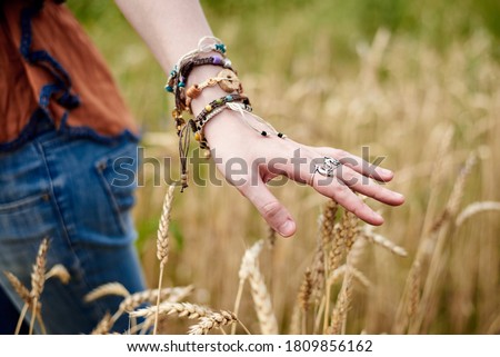 Close-up picture of woman's hand with colorful bracelets and silver rings, touching wheat stalks. Hippie girl standing in the middle of field. Eco tourism concept. Nature protection.