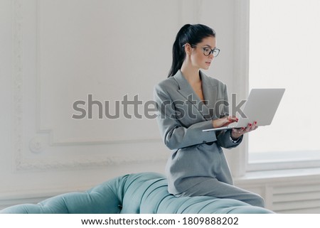 Photo of brunette executive worker poses in home office near sofa, holds laptop computer, makes financial report, wears optical glasses for vision correction, grey elegant suit, surfs internet