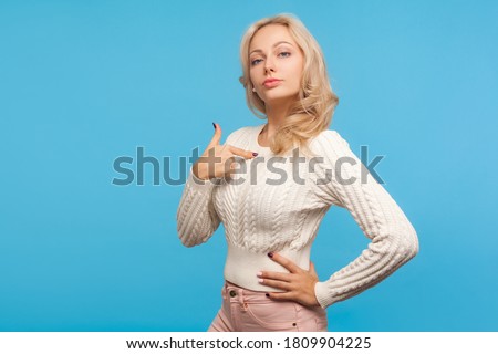 Self confident arrogant woman with curly blond hair pointing finger on herself, bragging with achievements, egoism. Indoor studio shot isolated on blue background Royalty-Free Stock Photo #1809904225