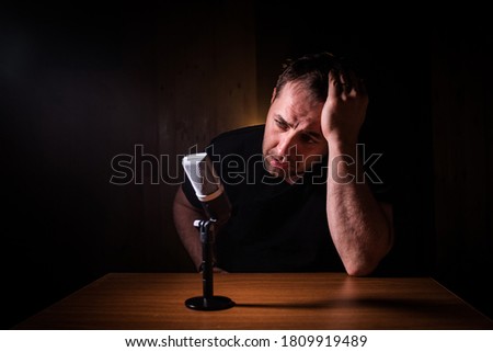 Caucasian man news broadcaster sat at a desk with microphone in dark room. Performer with microphone. Male musician singing into a microphone. Shot on a black background. Dark atmosphere