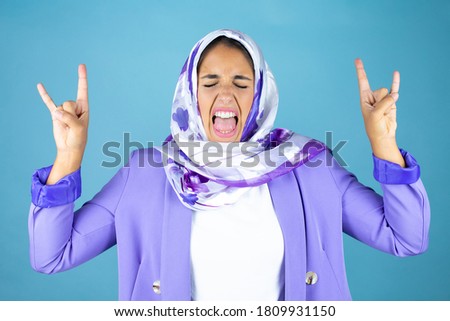 Young beautiful arab woman wearing islamic hijab over isolated blue background shouting with crazy expression doing rock symbol with hands up