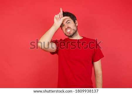 Young handsome caucasian man wearing t-shirt over isolated red background making fun of people with fingers on forehead doing loser gesture mocking and insulting.