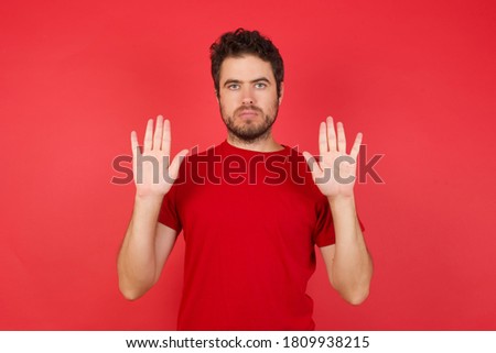 Serious Young handsome caucasian man wearing t-shirt over isolated red background pulls palms towards camera, makes stop gesture, asks to control your emotions and not be nervous