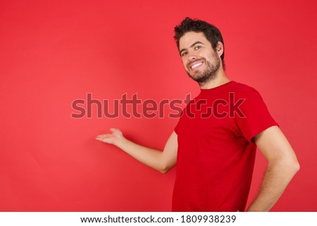 Young handsome caucasian man wearing t-shirt over isolated red background feeling happy and cheerful, smiling and welcoming you, inviting you in with a friendly gesture 