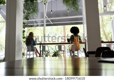 Two diverse young women sitting apart at cafe table, African and Caucasian ladies working or studying, dining in cafeteria avoiding communication, social distance safety concept, back view. Royalty-Free Stock Photo #1809975610