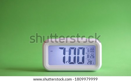 Digital alarm clock on table with green background. Displaying 7:00 am in the morning.