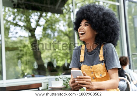 Cheerful African American hipster woman using phone, laughing, sitting at outdoor cafe table. Smiling happy mixed race black lady with afro hair having fun or feeling excited holding smart phone. Royalty-Free Stock Photo #1809988582