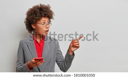 Stunned Afro American female enterpreneur points sideways right copy space, recommends product, sees astonishing thing, dressed in formal clothing, uses mobile phone, isolated on grey background