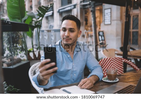 Young concentrated ethnic male in casual clothing trying to get internet signal while working on laptop remotely in cozy street cafe