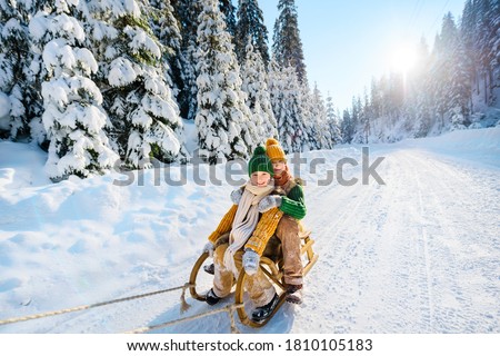 Two cheerful happy children ride on a wooden retro sled against the backdrop of a beautiful winter landscape with snow-covered fir trees. Sunny winter day.