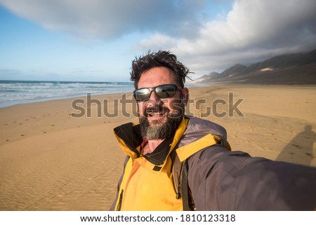 Happy wild lifestyle people concept with cheerful handsome adult man in wild beach with mopuntains in background - alternative summer vacation with adventure taste - caucasian male selfie