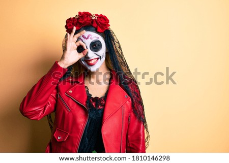 Woman wearing day of the dead costume over yellow smiling happy doing ok sign with hand on eye looking through fingers 