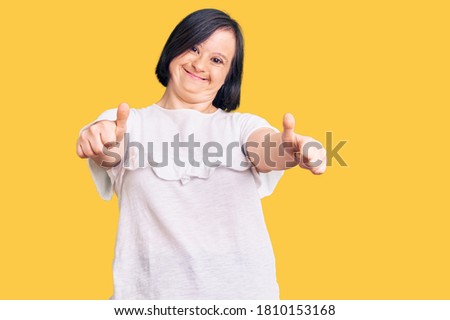 Brunette woman with down syndrome wearing casual white tshirt approving doing positive gesture with hand, thumbs up smiling and happy for success. winner gesture. 