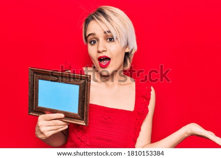Young beautiful blonde woman holding empty frame celebrating achievement with happy smile and winner expression with raised hand 