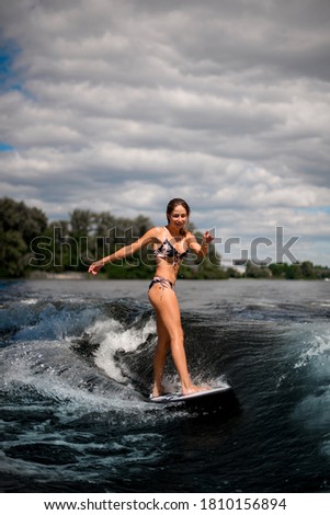 sporty smiling girl in bikini swimsuit stand on surfboard and vigorously rides on wave against the background of the bridge