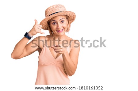 Young blonde woman wearing summer hat smiling doing talking on the telephone gesture and pointing to you. call me. 
