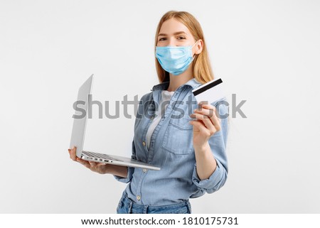young woman in a medical mask on her face, with a laptop and a plastic credit card in hand, on a white background