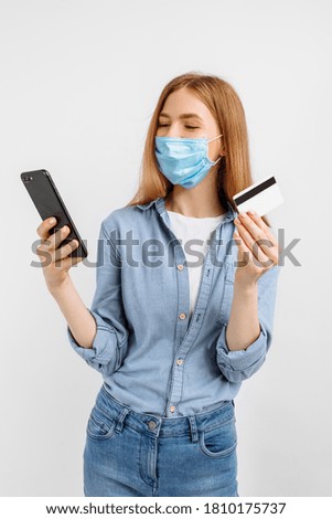 Happy young woman in a medical mask on her face, showing a plastic credit card and using a mobile phone, on a white background