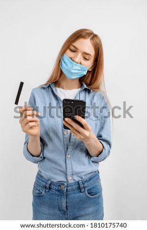 young woman in a medical mask on her face, showing a plastic credit card and using a mobile phone, on a white background