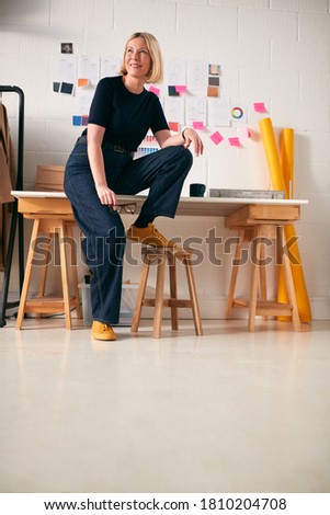 Portrait Of Confident Mature Businesswoman Sitting On Desk In Start Up Fashion Business