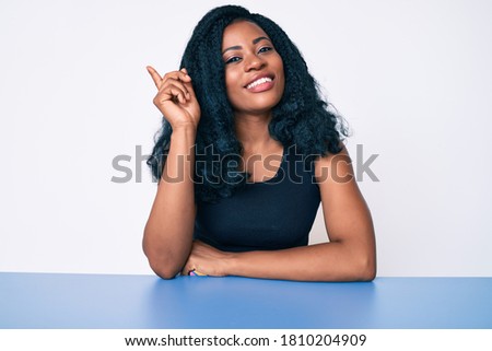 Beautiful african woman wearing casual clothes sitting on the table with a big smile on face, pointing with hand and finger to the side looking at the camera. 