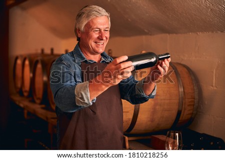 Joyful winemaker in apron holding bottle of alcoholic drink and smiling while spending time in wine storage Royalty-Free Stock Photo #1810218256