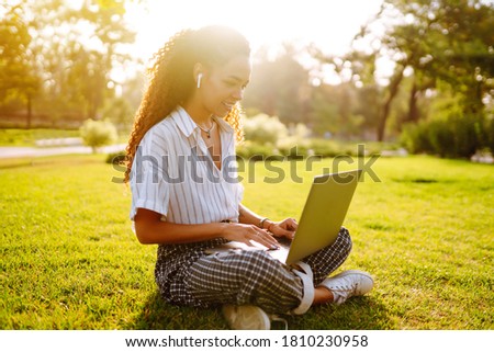 Portrait of pretty young woman sitting on green grass in park with  laptop. Happy woman, student, freelancer with wireless headphones calling on laptop, talk by webcam, video conference.
