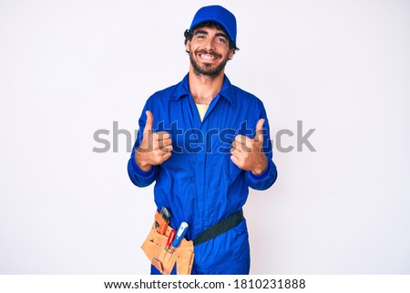 Handsome young man with curly hair and bear weaing handyman uniform success sign doing positive gesture with hand, thumbs up smiling and happy. cheerful expression and winner gesture. 