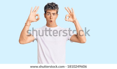 Young handsome man wearing casual white tshirt looking surprised and shocked doing ok approval symbol with fingers. crazy expression 