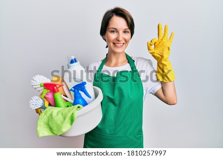 Young brunette woman with short hair wearing apron holding cleaning products smiling positive doing ok sign with hand and fingers. successful expression. 