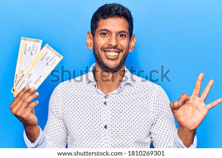 Young latin man holding boarding pass doing ok sign with fingers, smiling friendly gesturing excellent symbol 
