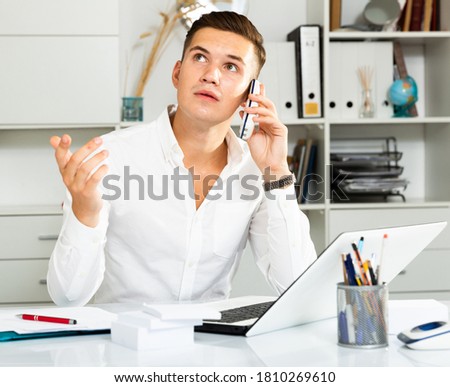 young guy in white shirt works at table with laptop and talking on phone in office