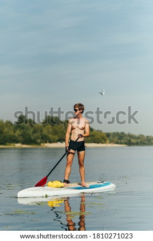 Portrait of a handsome young man with a sporty torso standing on the river on a sunny day on a sup board with an oar in his hands and resting, looking away. Vertical.