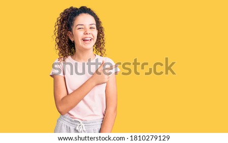 Beautiful kid girl with curly hair wearing casual clothes cheerful with a smile of face pointing with hand and finger up to the side with happy and natural expression on face 