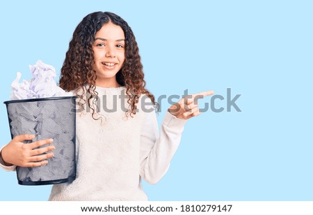 Beautiful kid girl with curly hair holding paper bin full of crumpled papers smiling happy pointing with hand and finger to the side 