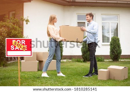Moving day. Young woman and her husband carrying carton box at house yard