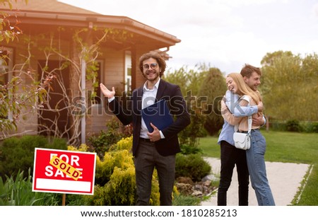Smiling house agent and happy couple hugging in front yard of their new property