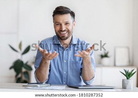 Successful handsome businessman sitting at desk in office, talking at camera, sharing his experience and ideas, free space Royalty-Free Stock Photo #1810287352