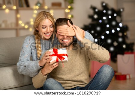 Beautiful woman surprising her husband with Christmas gift at home, closing his eyes