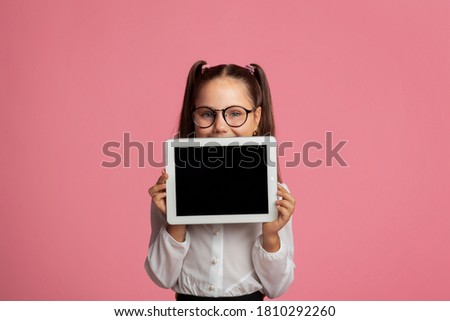 Education and distance learning for kids. Cheerful girl in glasses shows tablet with blank screen, isolated on pink background, studio shoot