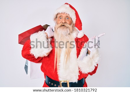 Old senior man with grey hair and long beard wearing santa claus costume holding shopping bags smiling happy pointing with hand and finger to the side 