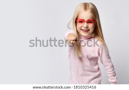 Cheerful blonde kid girl in stylish smoked heart-shaped sunglasses and turtleneck shirt sweater pointing finger at camera gesturing over gray background with free copy space