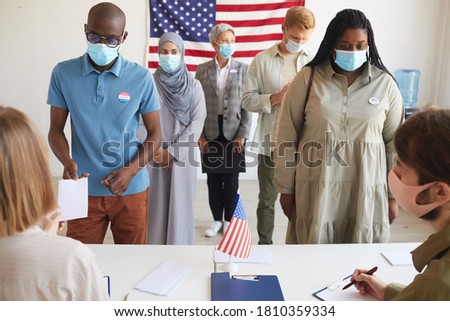 Front view at multi-ethnic group of people standing in row and wearing masks at polling station on election day, focus on two African-American people registering for voting Royalty-Free Stock Photo #1810359334