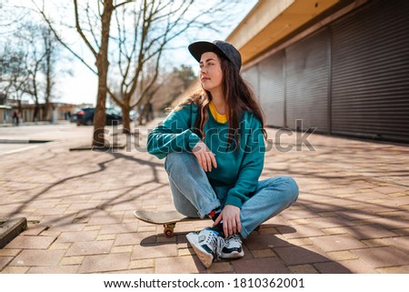 A young beautiful Caucasian woman sits relaxed on a skateboard and looks away. In the background, a street with trees. Concept of sports lifestyle and street culture