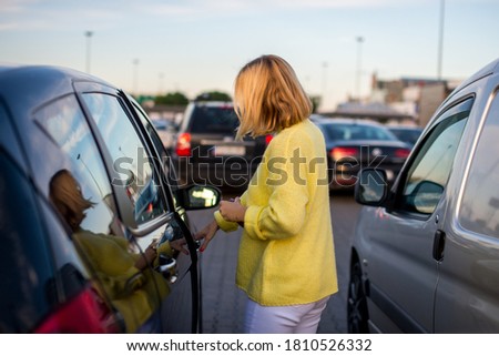 A young woman with a coronavirus mask in a yellow sweater gets into her car. The car is parked in the parking. There is bokeh in the background. Coronavirus concept. Life with covid-19. 