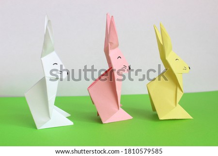 Pink, white and yellow origami rabbits on white and green background. Easter bunny on the grass. Minimalistic illustration. Paper craft.