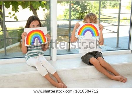 Little children holding rainbow paintings near window indoors. Stay at home concept