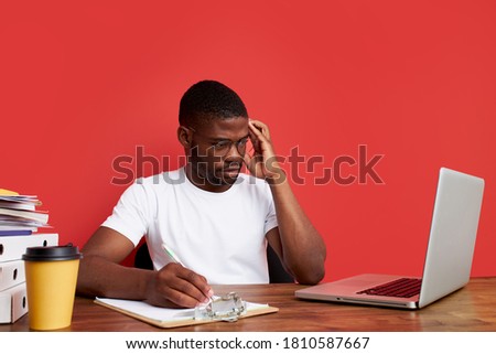 overworked unhappy and frustrated young african man in stress sitting at office desk, he has a lot of tasks in work, deadline, isolated red background