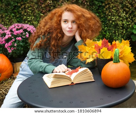 Beautiful young woman reading a romantic book in a cozy warm atmosphere enjoying the autumn mood. Rustic iron table with yellow mple flowers and orange pumpkin. redhead happy girl looking at far away